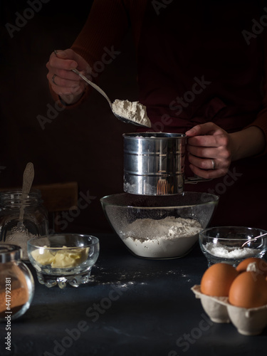 Cooking a cake. Put the flour in a sieve with a spoon. On the table are ingredients for making a pie, kitchen utensils. In the background is a girl in an apron. Dark background