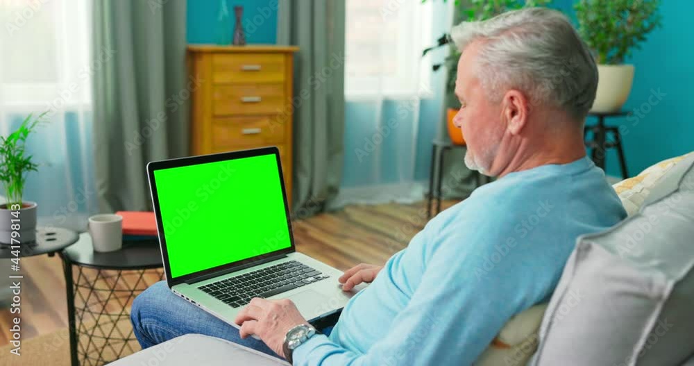 Older Man at Home Sitting on a Couch Works on a Laptop Computer with Green Mock-up Screen. Man Using Computer, Browsing through Internet, Watching Content, Chatting in Social Networks with Friends. Older Man at Home Sitting on a Couch Works on a Laptop Computer with Green Mock-up Screen. Man Using Computer, Browsing through Internet, Watching Content, Chatting in Social Networks with Friends.
