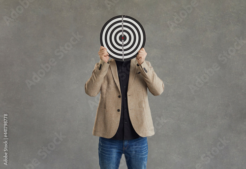 Unknown businessman hid his face behind a darts board while standing on a gray background. Concept of setting business goals, finding the target audience, achieving goals and success