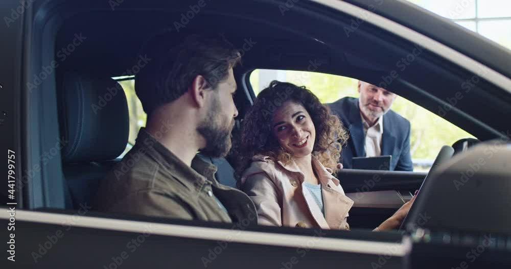 Husband and wife talking while sitting in dream car at automobile dealership. Couple of buyers choosing car for purchase and testing it. Bearded salesnmanager car agent in suit help the clients Husband and wife talking while sitting in dream car at automobile dealership. Couple of buyers choosing car for purchase and testing it. Bearded salesnmanager car agent in suit help the clients