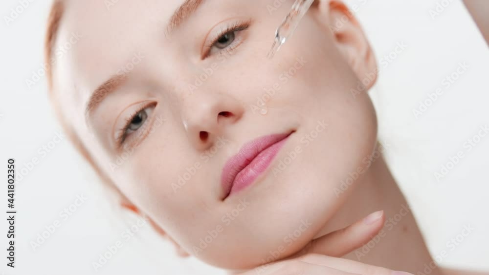 Attractive young slim red head European woman drips skin care serum from dropper on her cheek smiling for the camera on white background | Acne prevention concept Attractive young slim red head European woman drips skin care serum from dropper on her cheek smiling for the camera on white background | Acne prevention concept