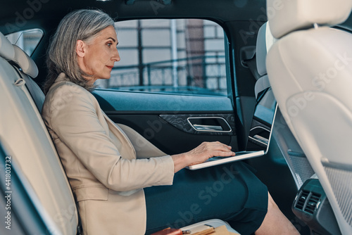 Confident mature woman in smart casual wear using laptop while sitting on the back car seat © gstockstudio