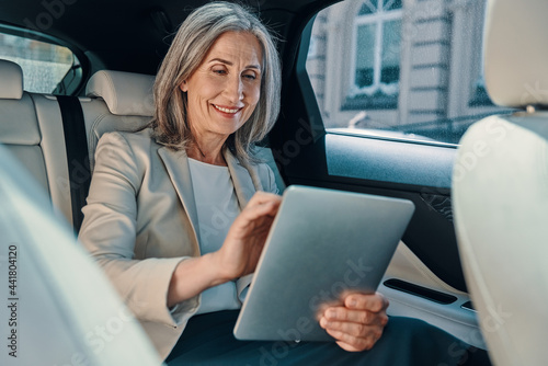 Mature beautiful woman in smart casual wear using digital tablet while sitting on the back car seat © gstockstudio