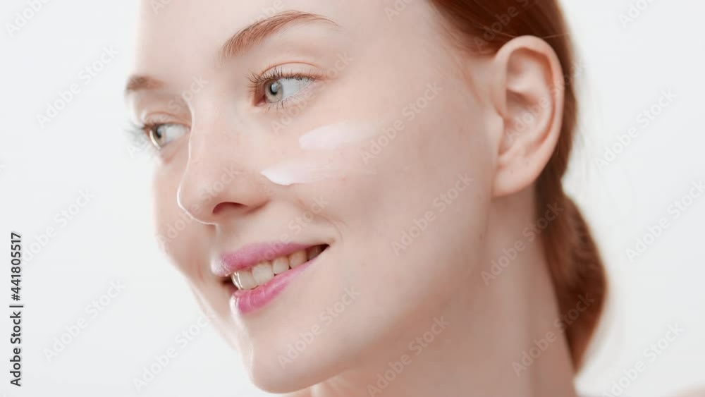 Pretty slim European girl with long orange hair applies moisturizer on her cheek smiling wide on white background | Moisturizer commercial Pretty slim European girl with long orange hair applies moisturizer on her cheek smiling wide on white background | Moisturizer commercial