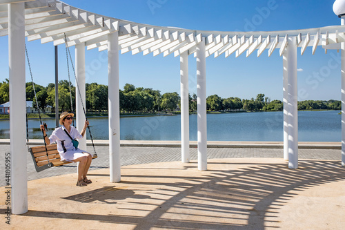 A girl in a white shirt and a straw hat is swinging on a swing in the park under a white canopy that protects from the sun.