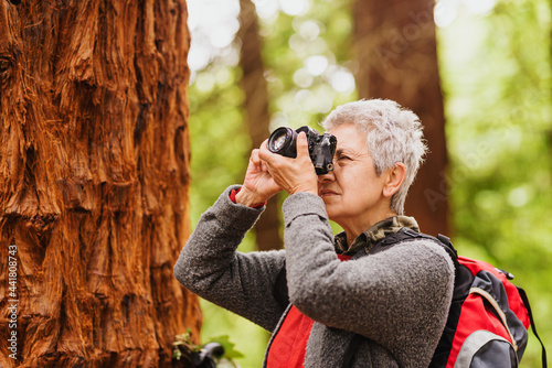 elderly woman with gray hair and backpack taking pictures with an old camera during a hike in the woods.