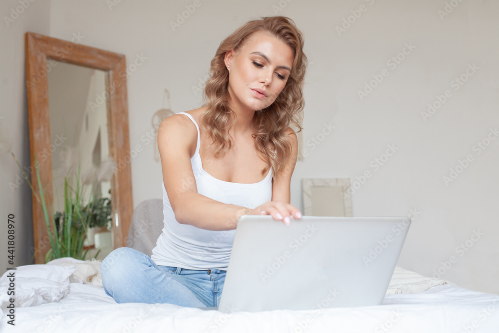 Young casual beautiful woman ending work on a laptop sitting on the bed in the house Young casual beautiful woman ending work on a laptop sitting on the bed in the house