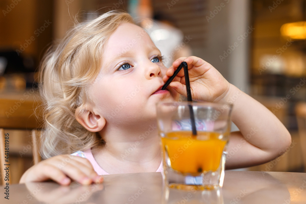 A beautiful blonde baby girl is sitting at a table in a cafe and drinking juice through a straw. A beautiful blonde baby girl is sitting at a table in a cafe and drinking juice through a straw.