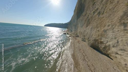 Rapid fast movement fpv sport drone over natural beach sea coast near high mountain rock at summer sunny day. Aerial shot view flying at seashore with calmness waving water and clear blue sunny sky