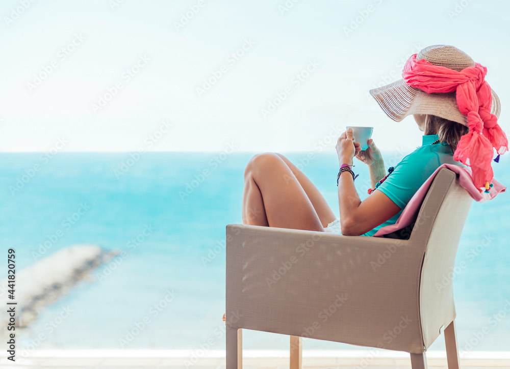 A middle-aged woman sitting in an armchair contemplates the blue sea while drinking a coffee on the terrace of a hotel. Sitges beach, Barcelona.