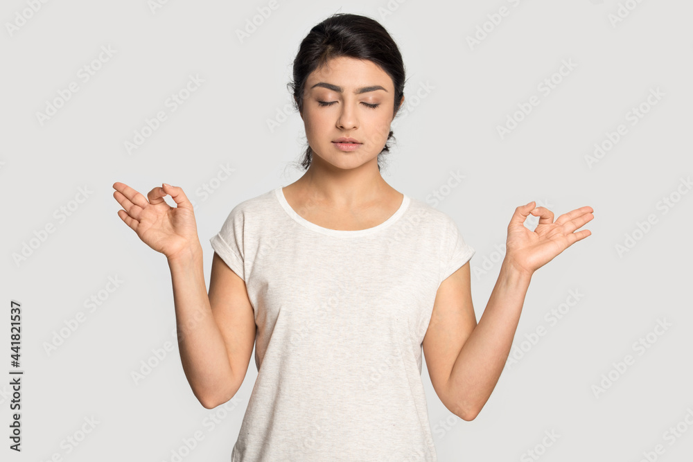 Calm Indian young woman isolated on grey studio background practice yoga breathe fresh air. Peaceful millennial mixed race female with mudra hands meditate relieve negative emotion. Peace concept. Calm Indian young woman isolated on grey studio background practice yoga breathe fresh air. Peaceful millennial mixed race female with mudra hands meditate relieve negative emotion. Peace concept.