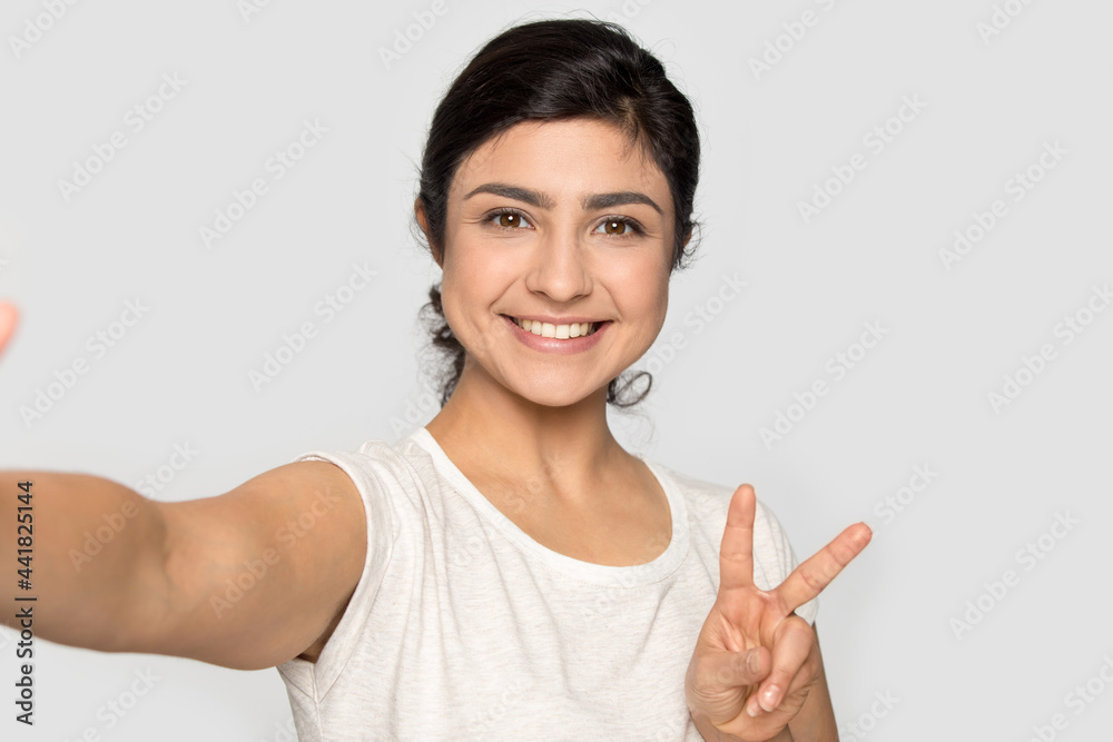 Smiling Indian young woman isolated on grey studio background look at camera make self-portrait picture. Happy millennial mixed race female pose with victory hand gesture take selfie on modern gadget. Smiling Indian young woman isolated on grey studio background look at camera make self-portrait picture. Happy millennial mixed race female pose with victory hand gesture take selfie on modern gadget.