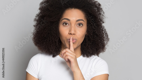 Portrait of young African American woman isolated on grey studio background keep finger at lips tell secret. Millennial mixed race female share secret good sale deal or promotion discount to clients.