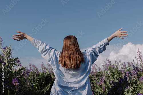 Unrecognizable woman with arms outstretched standing alone among field of purple flowers in front of blue sky enjoying of sunlight. Girl from the back. Summer outfit. Having fun outdoors.
