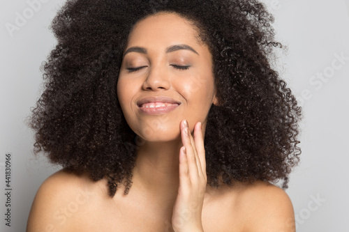 Closeup of happy millennial African American woman isolated on grey background touch healthy glowing skein after face treatment. Smiling young biracial female satisfied with skincare procedures.