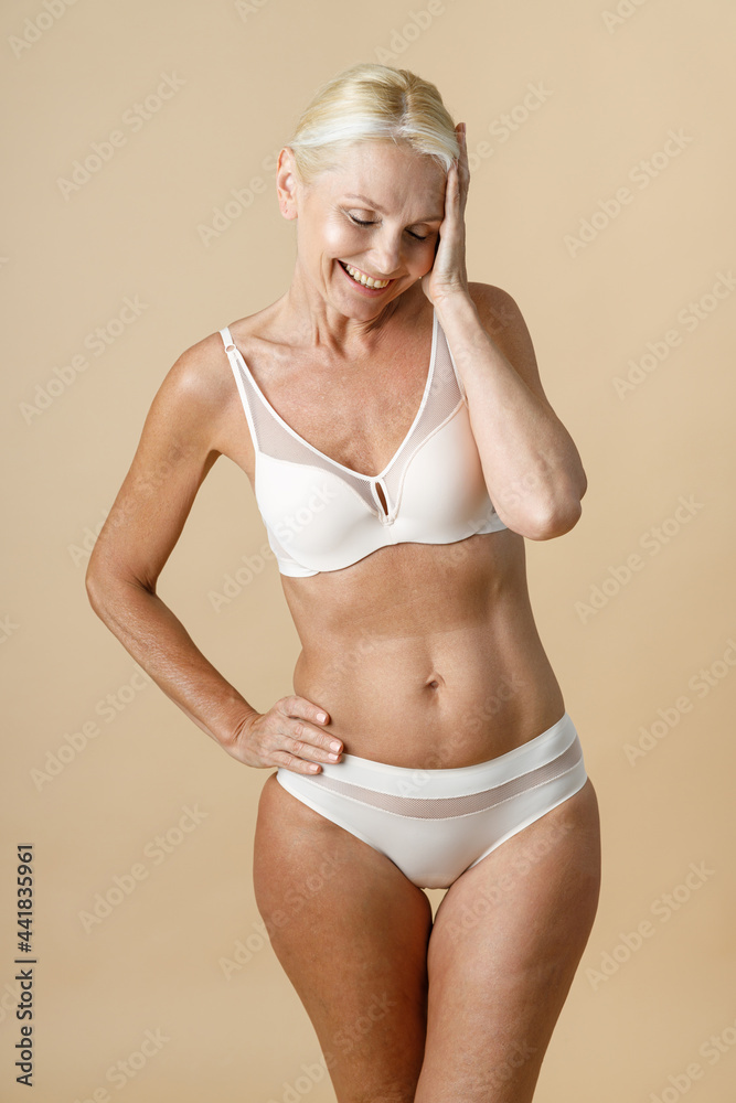 Charming mature woman in underwear with fit perfect body smiling and  touching her head, standing with eyes closed isolated over beige background  Photos | Adobe Stock
