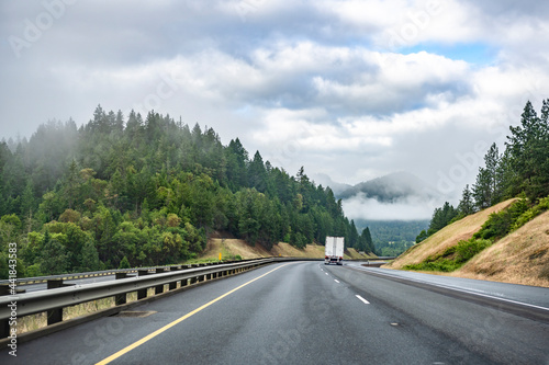 Big rig semi truck transporting cargo in semi trailer running on the scenic divided highway road between green hills with forest