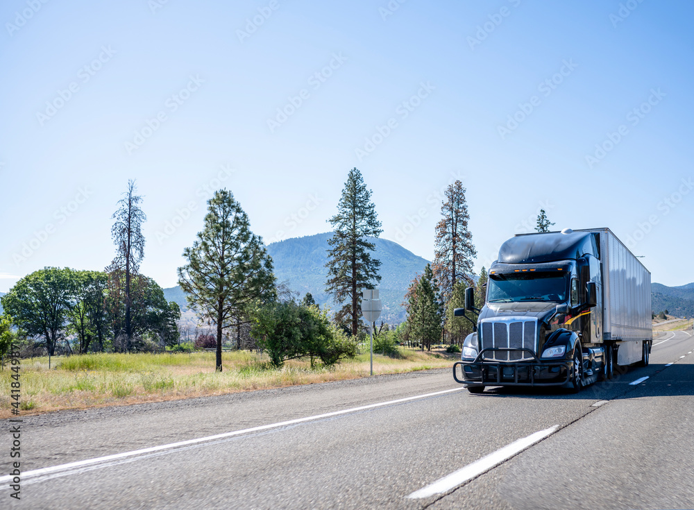 Black stylish big rig bonnet industrial semi truck with dry van semi trailer driving on the highway road with trees and mountain on background