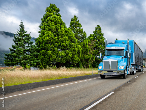 Blue big rig classic semi truck for long haul transporting cargo in covered semi trailer with front wall spoiler driving on the interstate highway