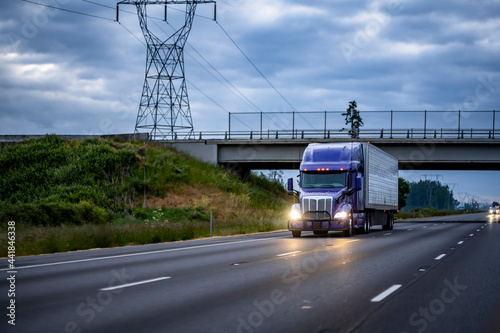 Bright blue big rig semi truck transporting cargo in refrigerated semi trailer running under the bridge on the wide highway road at the night time