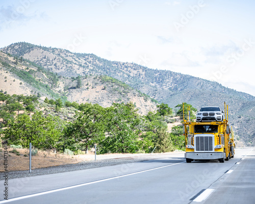Bright yellow professional industrial big rig car hauler semi truck transporting cars on the modular semi trailer running on the road with bald mountains of summer California on the background