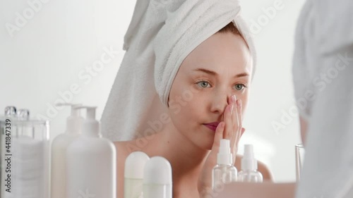Pretty fit European young woman wrapped in a towel sits down in front of the mirror in bathroom and finds some skin problems disappointing on white background | Skincare concept