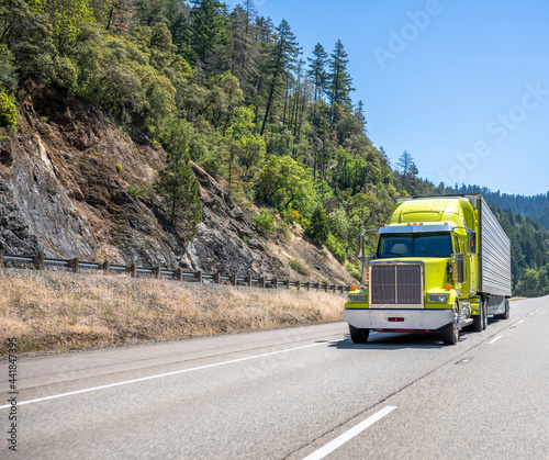 Built with the latest aerodynamic advancements big rig lime semi truck transporting cargo in refrigerated semi trailer running on the mountain road with rocks and trees
