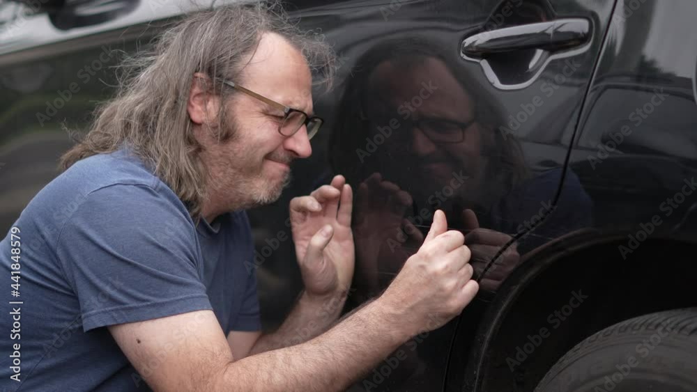 A middle-age man looking closely to the paint of his car and being upset about the scratches