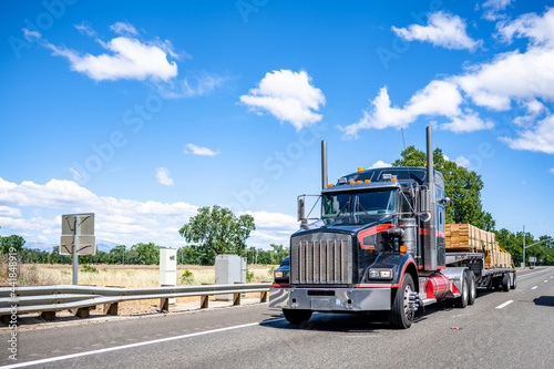 Classic style black big rig semi truck with red accents transporting lumber wood on the step down semi trailer driving on the highway road
