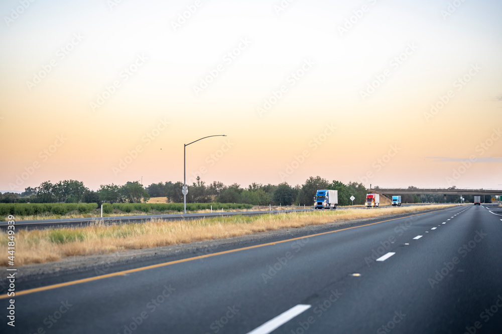 Fototapeta premium Convoy of loaded big rigs semi trucks with semi trailers running on the evening divided highway road