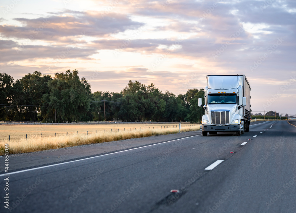 Industrial bonnet American big rig semi truck with covered dry van semi trailer driving on the evening twilight highway road with colorful clouds