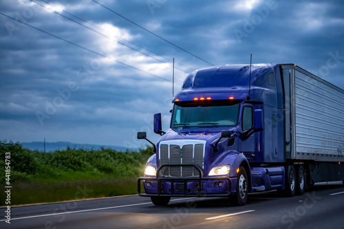 Stylish blue big rig semi truck transporting frozen cargo in refrigerator semi trailer running on the evening wide highway road with storm clouds