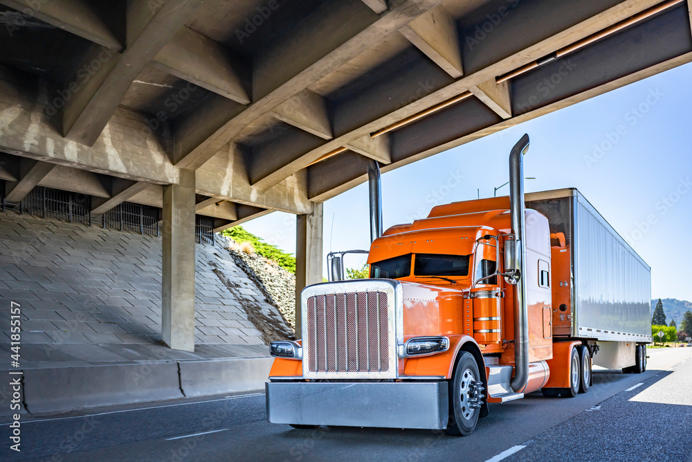 Stylish orange classic rig semi truck with chrome accessories and exhaust pipes transporting cargo van low profile semi trailer running on the highway road under the bridge Photos