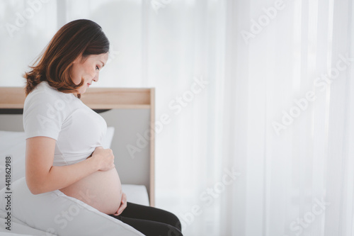 Asian beautiful and happy woman in pregnancy sitting beside the bed in bedroom. Free Download Asian beautiful and happy woman in pregnancy sitting beside the bed in bedroom. - GFXLoad