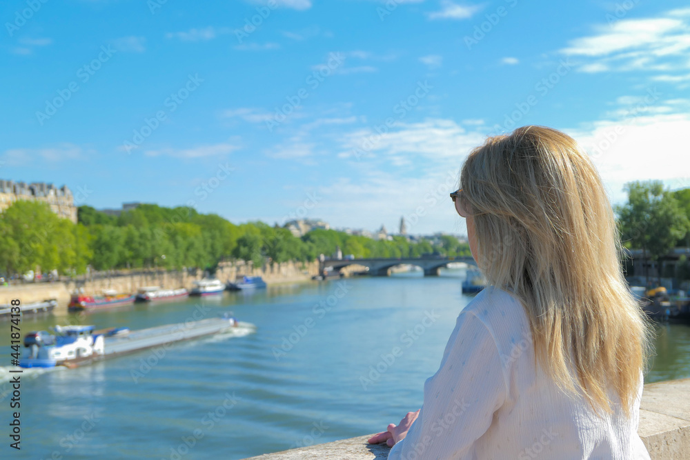 Blonde woman from behind on a bridge in Paris looking at the Seine river in summer. Barge blurred deliberately in the background.