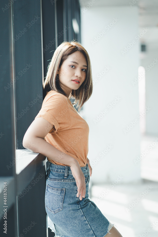 Stylish Asian girl wearing orange t-shirt and jean skirt posing in empty room, urban clothing style. Street photography. Stylish Asian girl wearing orange t-shirt and jean skirt posing in empty room, urban clothing style. Street photography.