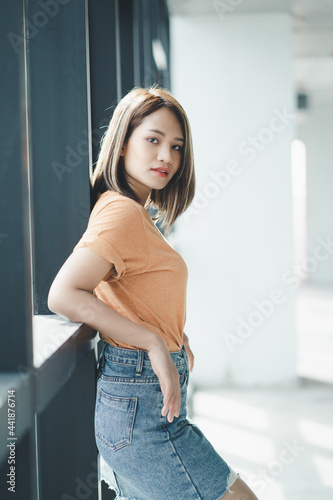 Stylish Asian girl wearing orange t-shirt and jean skirt posing in empty room, urban clothing style. Street photography.