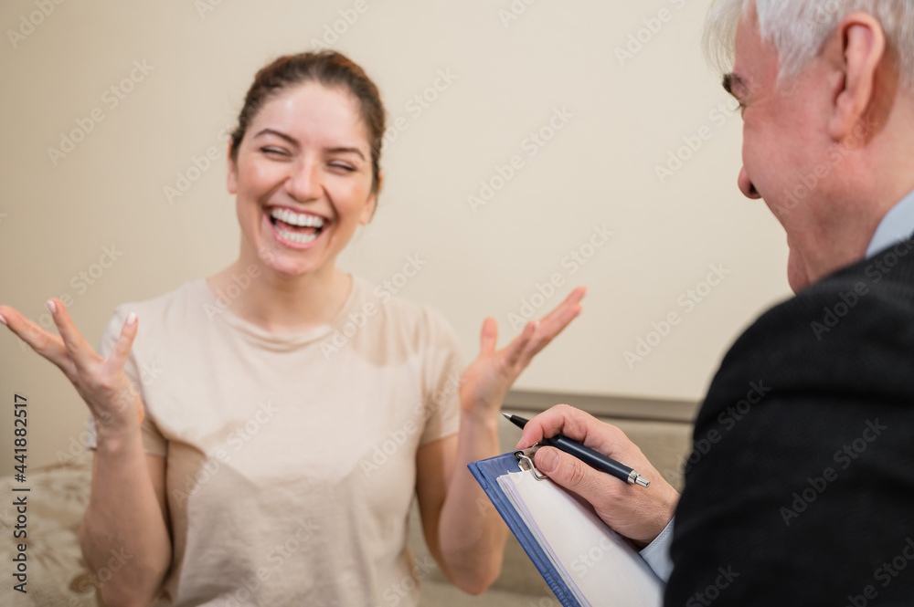 Fototapeta premium Mature gray-haired male psychotherapist with clipboard in session with female patient. Smiling caucasian woman at a psychologist appointment.