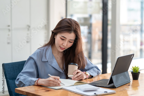 Asian businesswoman sitting at the office holding a cup of coffee and taking notes using a pen tablet placed at the office.