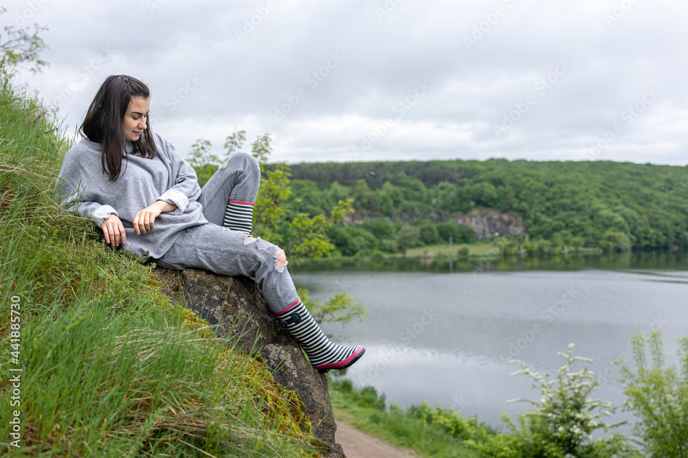 A girl in rubber boots sits on a stone near a lake in the forest.