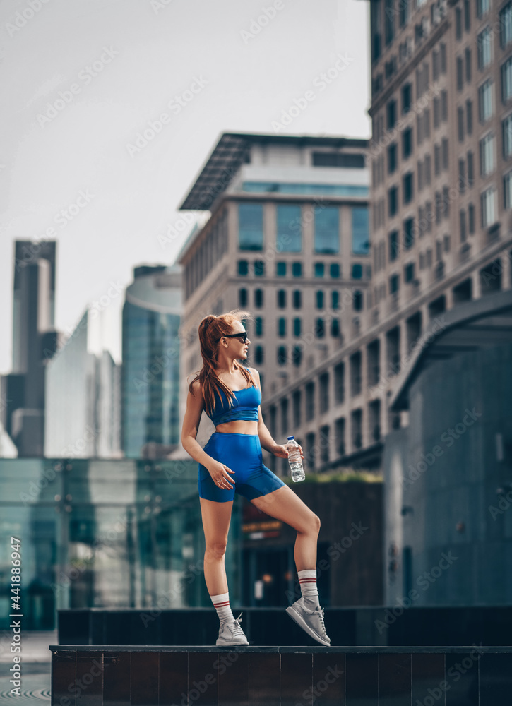 Young strong athletic red-haired woman in sportswear, runner stands on city street holding bottle of water in hand and looks aside.