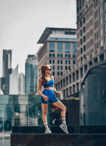 Young strong athletic red-haired woman in sportswear, runner stands on city street holding bottle of water in hand and looks aside.