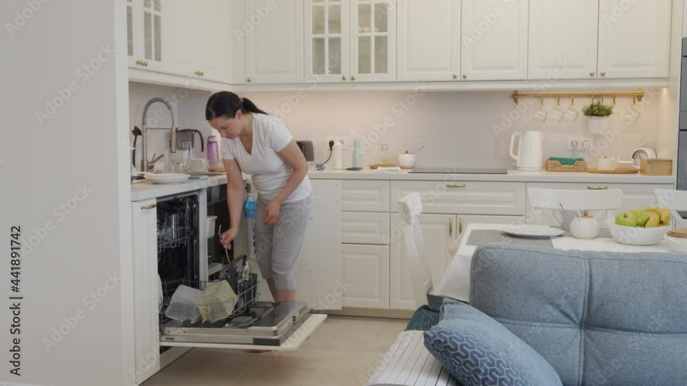 Housewife doing household chores cleaning kitchen, woman putting dishes into dishwasher, modern white apartment interior with open floor plan. High quality 4k footage Housewife doing household chores cleaning kitchen, woman putting dishes into dishwasher, modern white apartment interior with open floor plan. High quality 4k footage