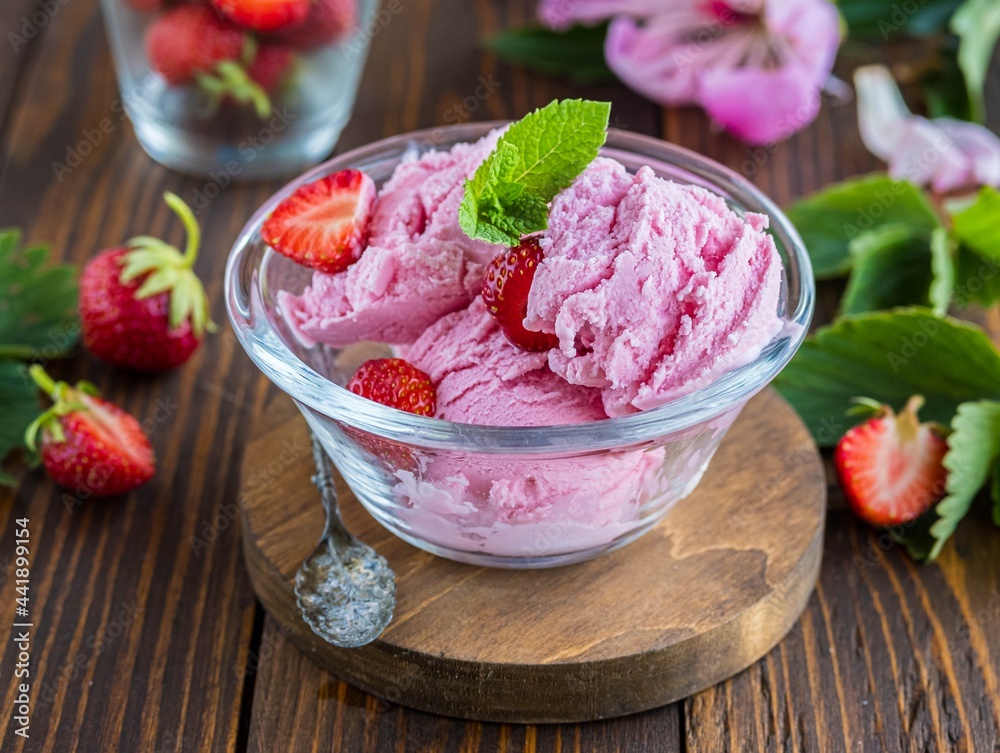 Three scoops of homemade strawberry ice cream with fresh strawberries in a transparent bowl on a brown wooden background. Three scoops of homemade strawberry ice cream with fresh strawberries in a transparent bowl on a brown wooden background.