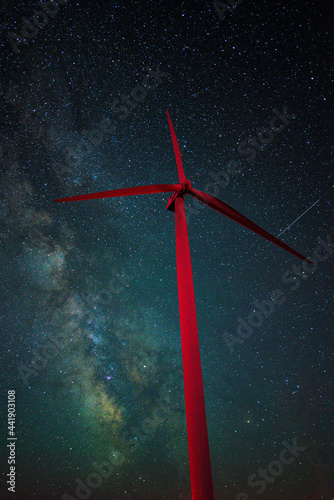 Astrophotography shot of a wind turbine and the Milky Way Galaxy as a shooting star passes behind the red wind turbine