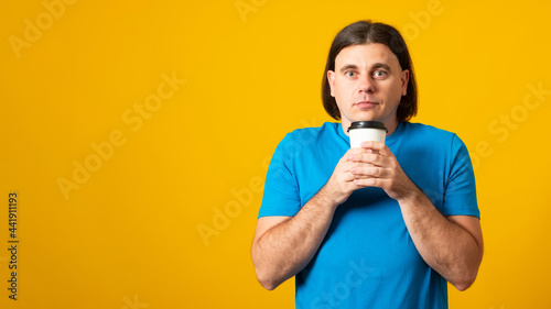 Banner. Handsome man with a fashionable hairstyle, drinking hot coffee a paper cup, trying to keep warm, wearing a blue T-shirt, on a yellow background with blank space for the text.