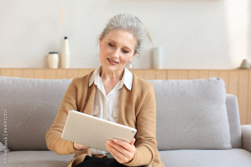 Cheerful middle aged woman sitting on sofa, using computer tablet apps, looking at screen,reading good news in social network, shopping or chatting online. Cheerful middle aged woman sitting on sofa, using computer tablet apps, looking at screen,reading good news in social network, shopping or chatting online.