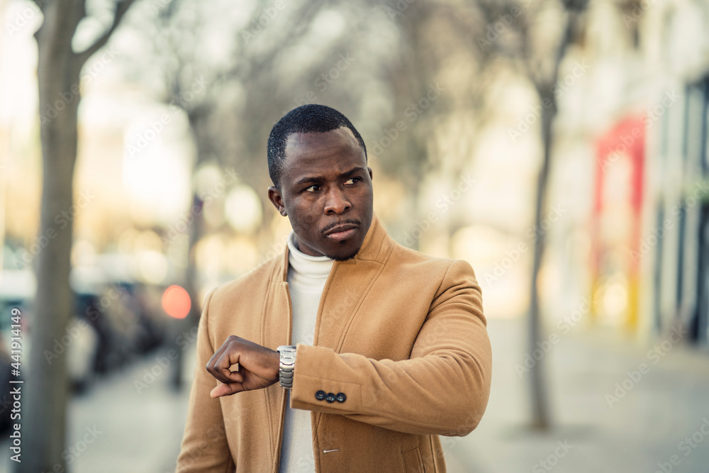 Serious black man checking time on wristwatch while strolling on ...