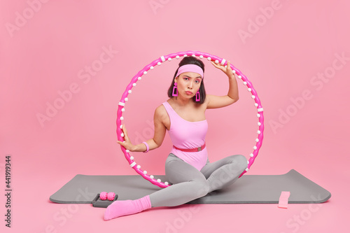 Slim fitness woman has training with hula hoop dressed in sportswear engaged in sport poses on mat looks attentively at camera isolated over pink background. Active lifestyle and home workout photo