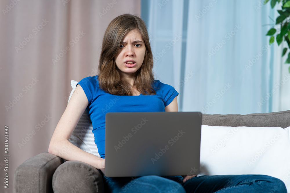 Fototapeta premium Portrait of scared stressed frighten teen girl, young afraid beautiful woman looking at screen of her laptop computer with fear, sitting it home on sofa or couch. Internet fraud concept.